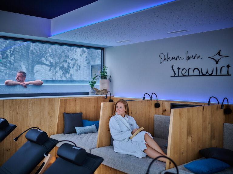 Eine Frau sitzt auf einer Liege im Ruheraum während ein Mann im Pool durch das Fenster schaut.